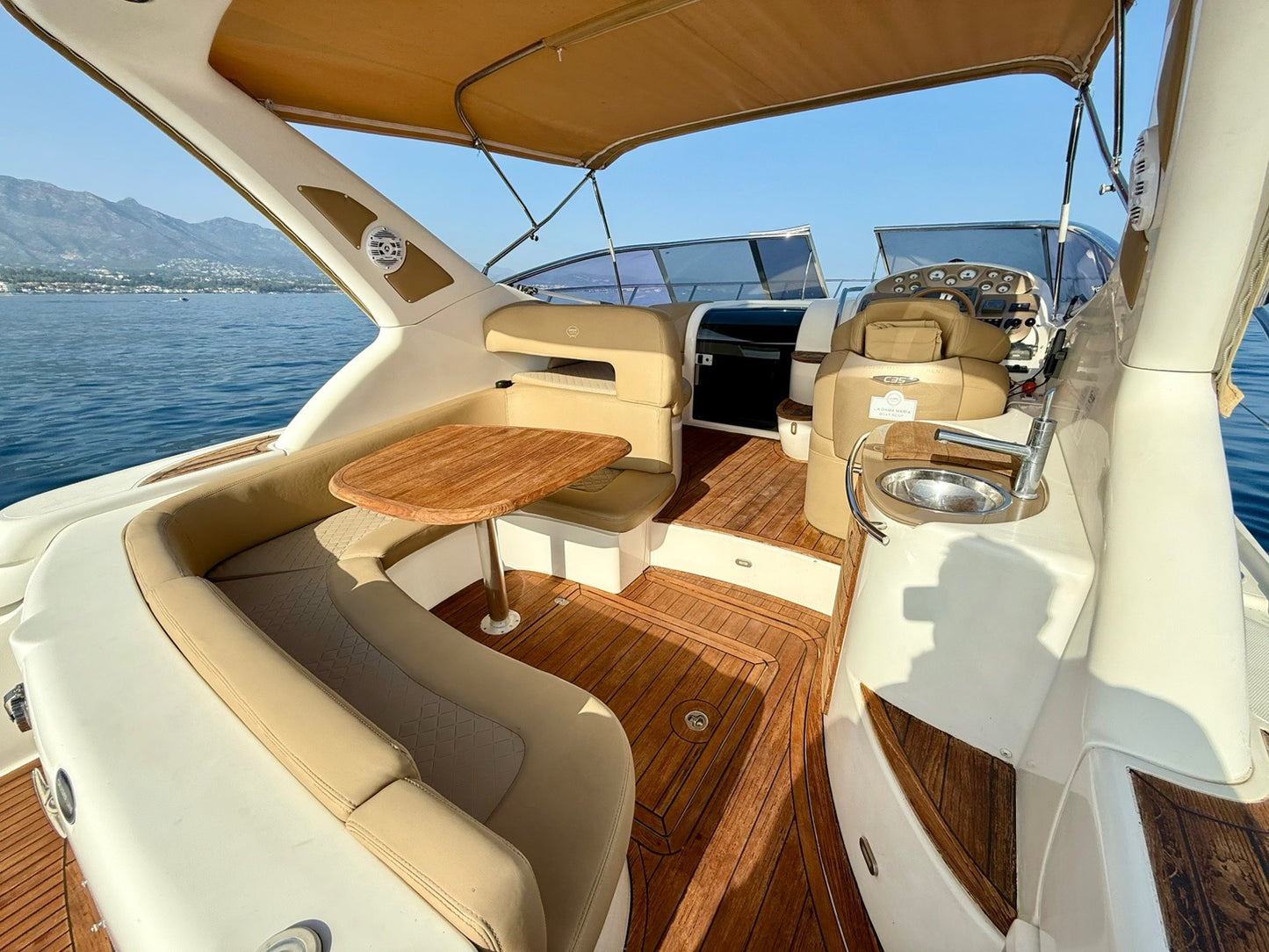 Interior of a luxurious yacht with wooden flooring and beige seating, surrounded by water and mountains.